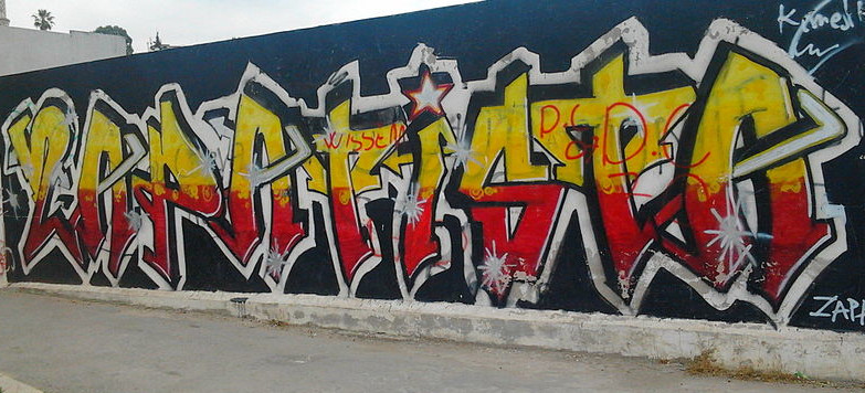Ein langes Wandgraffiti mit einem Verlauf von gelber in rote Schrift, der Schriftzug lautet ZAPATISTA.